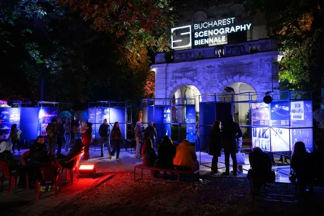 Oamenii stau pe scaune și bănci în fața unor panouri de expoziție iluminate în albastru și roșu, sub un semn luminat de eveniment.