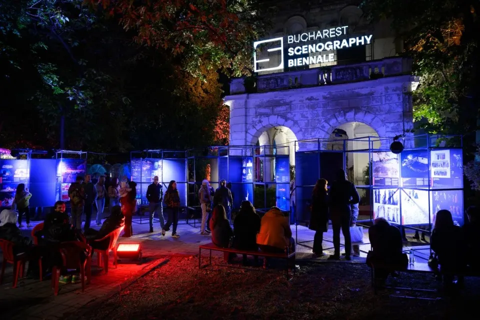 Oamenii stau pe scaune și bănci în fața unor panouri de expoziție iluminate în albastru și roșu, sub un semn luminat de eveniment.