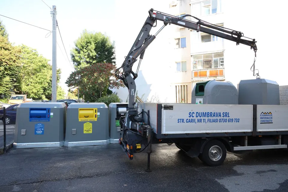 Un camion cu braț hidraulic descarcă containere de deșeuri de culoare gri lângă alte containere colorate pe un trotuar lângă un bloc de locuințe.