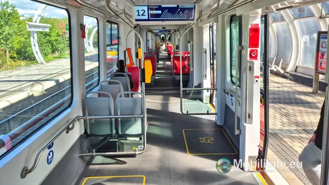 Interiorul unui tramvai cu scaune gri, uși deschise spre un peron și ferestre mari oferind o vedere spre vegetație și structuri metalice curbate.