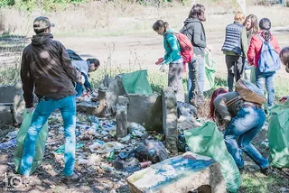 Un grup de persoane adună deșeuri în saci verzi pe un teren cu vegetație și resturi de beton.