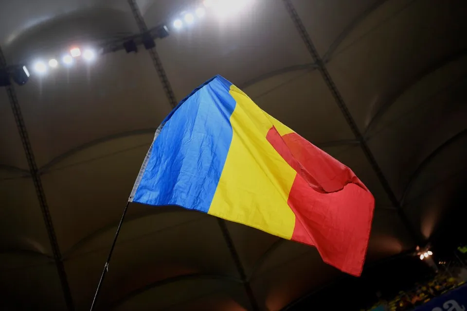 Un drapel tricolor cu benzi verticală albastră, galbenă și roșie este întins pe un stâlp sub un acoperiș întunecat iluminat de reflectoare.