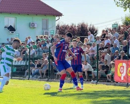 Cei doi jucători de fotbal în echipament albastru și roșu se află pe terenul de fotbal, în fața unor spectatori așezați pe tribune.