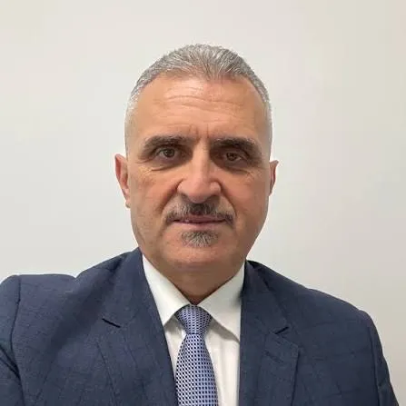 Un bărbat cu părul scurt și cărunt, purtând sacou albastru, cămașă albă și cravată cu model punctat, privește spre cameră pe un fundal neutru.