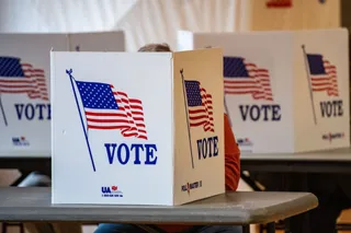 Mai multe cutii de votare albe cu imagini cu steaguri americane și textul VOTE sunt așezate pe mese într-un spațiu interior.