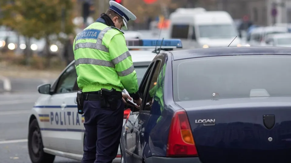 Un polițist în uniformă reflectorizantă stă lângă o mașină albastră și verifică un document lângă un vehicul de poliție alb.