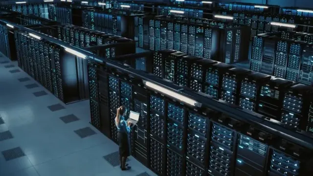 O persoană stă în picioare lângă rânduri de servere într-un centru de date, utilizând un laptop.