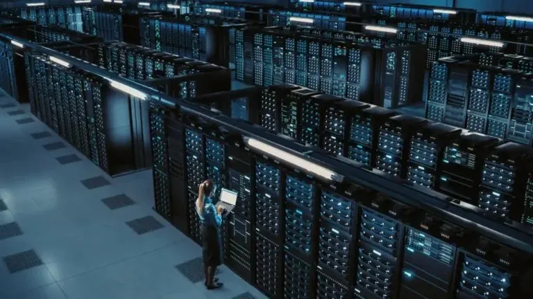 O persoană stă în picioare lângă rânduri de servere într-un centru de date, utilizând un laptop.