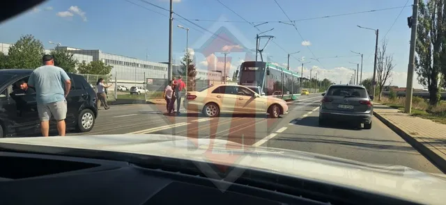 Un bărbat în șort și tricou albastru stă lângă o mașină neagră pe un drum asfaltat, lângă un autoturism galben și un tramvai.