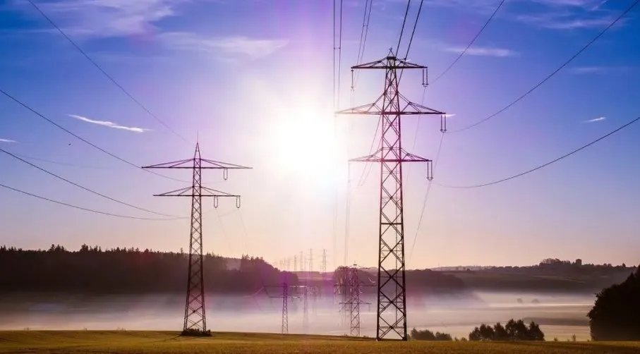 Două turnuri de înaltă tensiune stau pe un câmp verde sub un cer albastru cu soare strălucitor și linii electrice întinse.