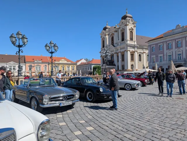 Mașini clasice sunt parcate pe o piață pavată în fața unei clădiri baroce sub un cer senin.