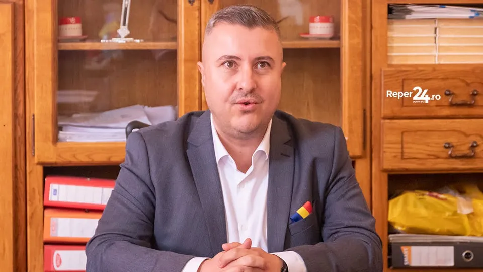 Un bărbat cu sacou gri și cămașă albă stă la o masă în fața unor dulapuri de lemn cu rafturi și dosare.