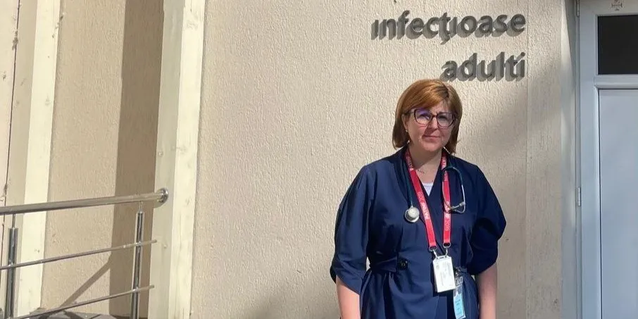 O femeie cu ochelari și un stetoscop purtând o uniformă albastră stă în fața unui perete pe care scrie text metalic.
