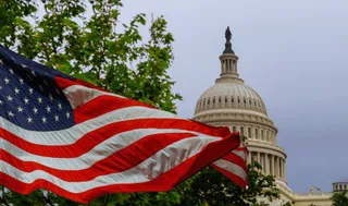 O steag american agitat în prim-plan apare în fața cupolei unei clădiri albe sub un cer gri.