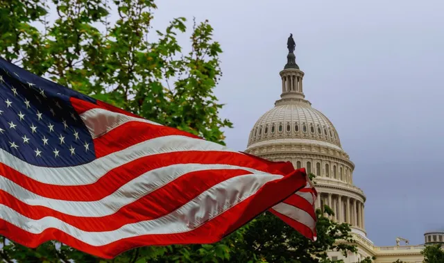 O steag american agitat în prim-plan apare în fața cupolei unei clădiri albe sub un cer gri.