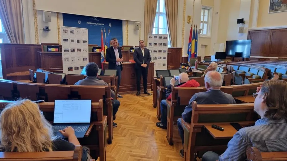 Un grup de bărbați stă în picioare în fața unor panouri de prezentare într-o sală de conferințe cu scaune de lemn și oameni așezați.