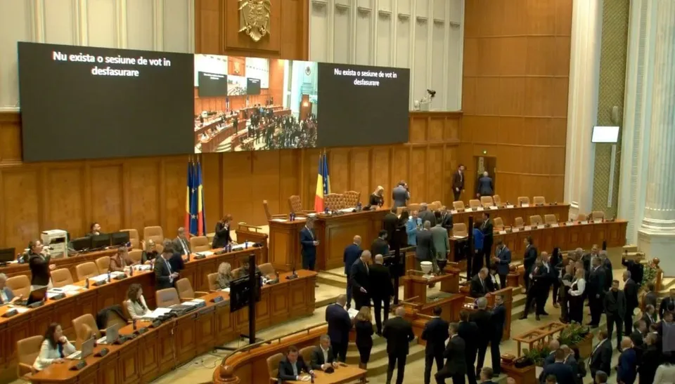 Oamenii stau în picioare în interiorul unei săli de ședințe cu mese de lemn și ecrane mari afișând text alb pe fundal negru.
