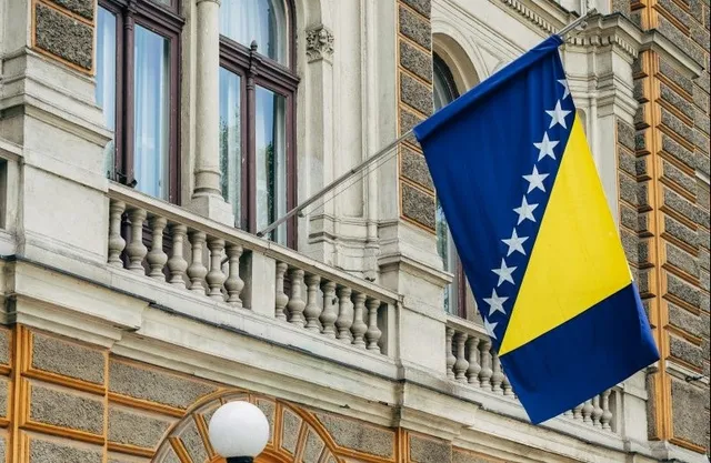 O steag al Bosniei și Herțegovine este arborat pe o fațadă de clădire cu balustradă și ferestre înalte.