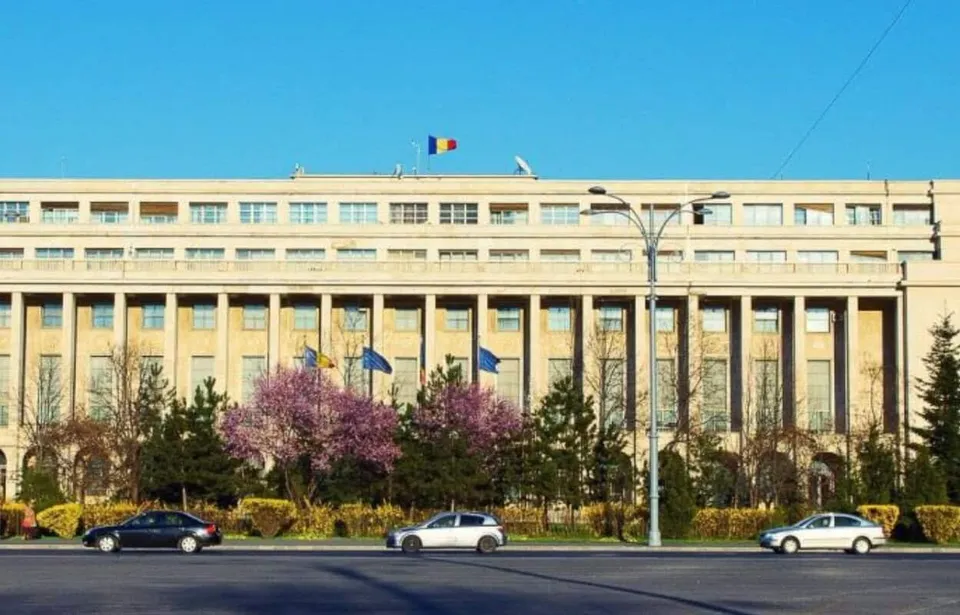 O clădire lungă cu multe ferestre este situată în spatele unor copaci, cu mașini parcate pe marginea unui drum în prim-plan.