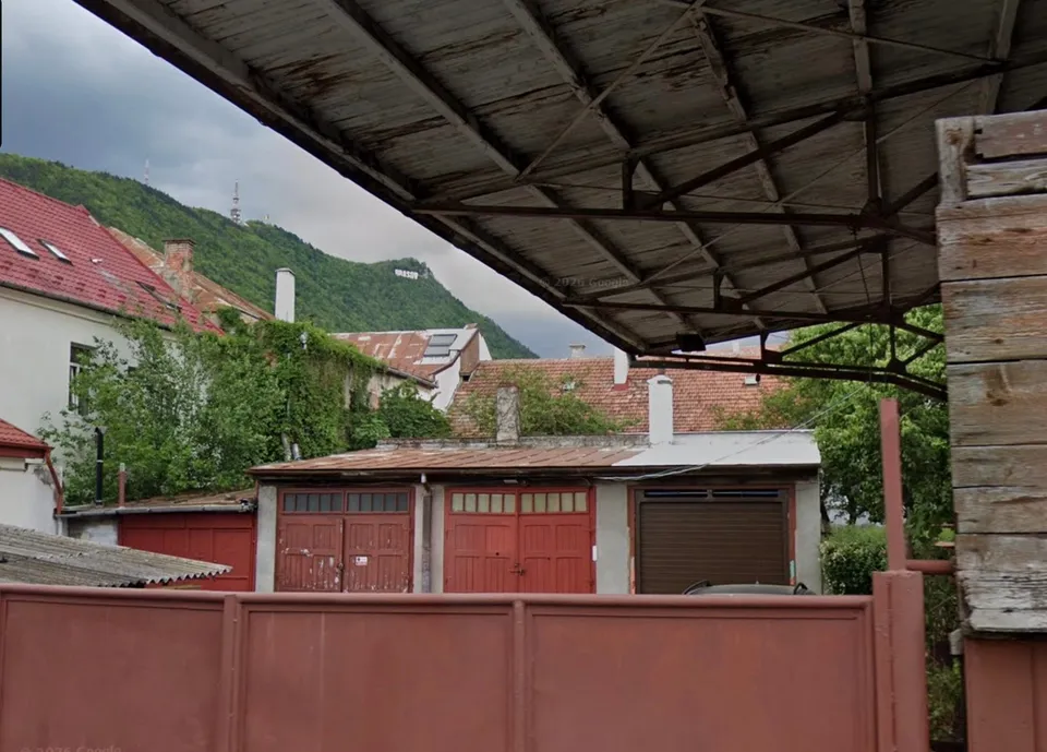 O serie de garaje cu uși metalice roșii și maronii sunt situate sub o structură de acoperiș din lemn și metal, lângă un deal verde.