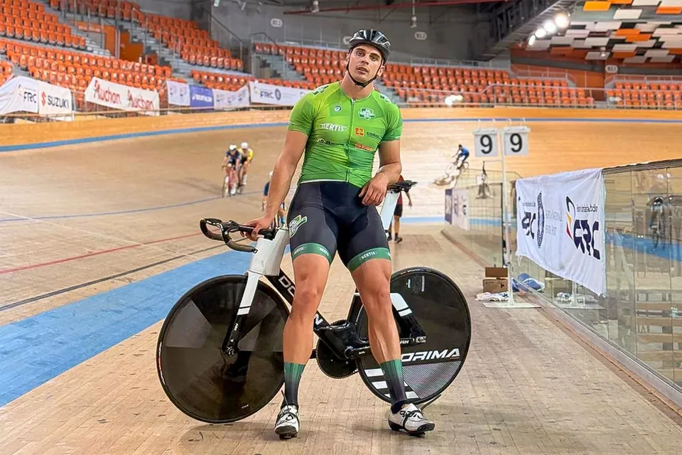 Un bărbat cu cască și echipament de ciclism stă în picioare lângă o bicicletă pe o pistă de ciclism interioară cu tribune portocalii în fundal.