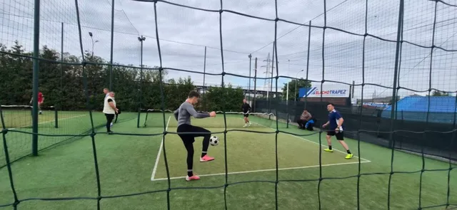 Câțiva oameni joacă fotbal pe un teren de sintetice verde, înconjurat de o plasă de protecție înaltă sub un cer înnorat.