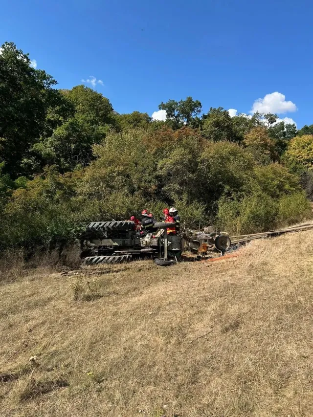 Un tractor este răsturnat pe o pante acoperită de iarbă uscată, în prezența unor persoane purtând echipament de protecție portocaliu și casci.