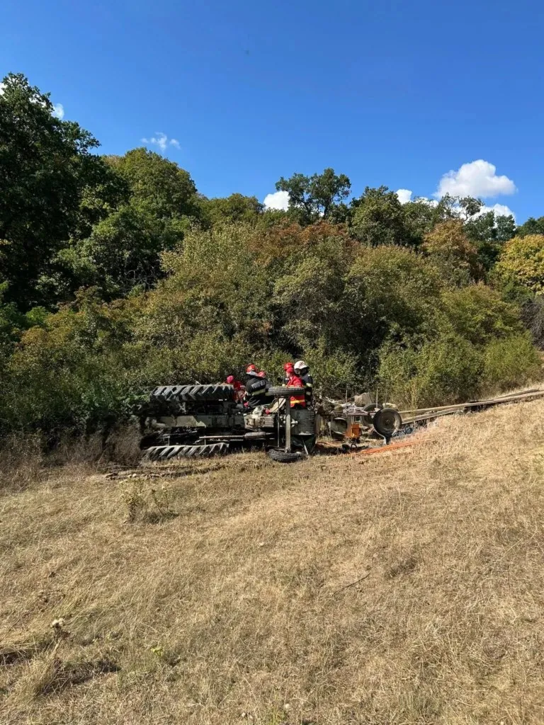 Un tractor este răsturnat pe o pante acoperită de iarbă uscată, în prezența unor persoane purtând echipament de protecție portocaliu și casci.
