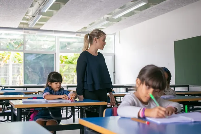 O profesoară stă în picioare lângă o elevă, în timp ce o altă elevă scrie la o bancă, într-o sală de clasă.