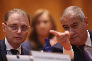 Doi bărbați în costum se află în primul plan, unul cu ochelari arăând în față și celălalt arăând cu degetul, cu o femeie în fundalul neclar.