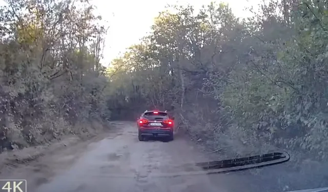Un autoturism roșu circulă pe un drum de pământ înconjurat de vegetație densă sub un cer luminos.