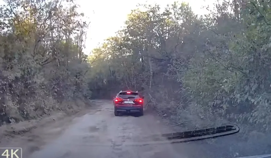 Un autoturism roșu circulă pe un drum de pământ înconjurat de vegetație densă sub un cer luminos.