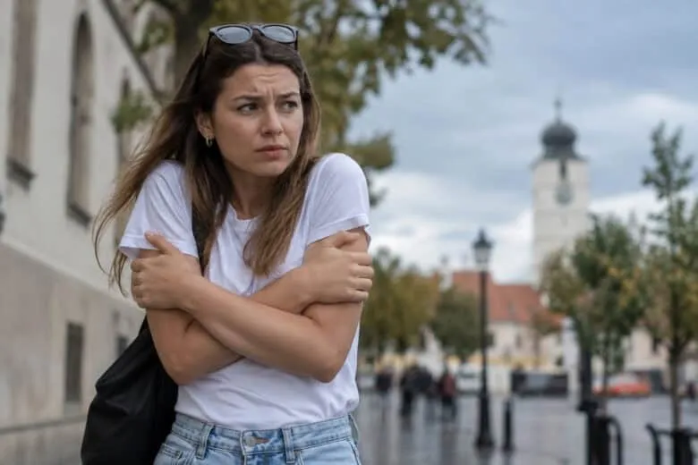 O femeie cu părul lung și tricou alb stă cu brațele încrucișate pe piept, privind spre lateral într-un decor urban cu clădiri și copaci.