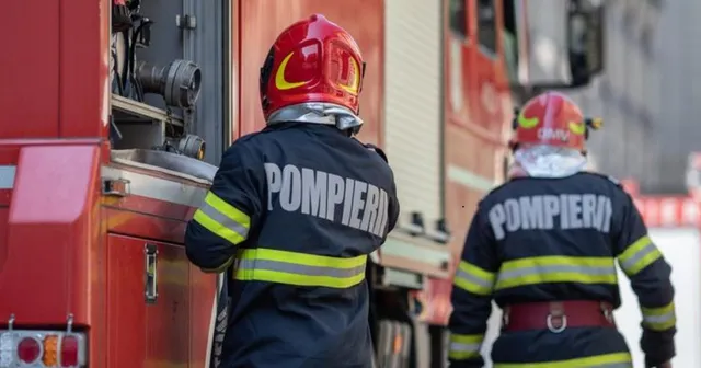Doi pompieri cu echipament de protecție și cască roșie stau lângă un autospecială de stingere a incendiilor.