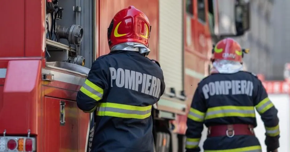 Doi pompieri cu echipament de protecție și cască roșie stau lângă un autospecială de stingere a incendiilor.