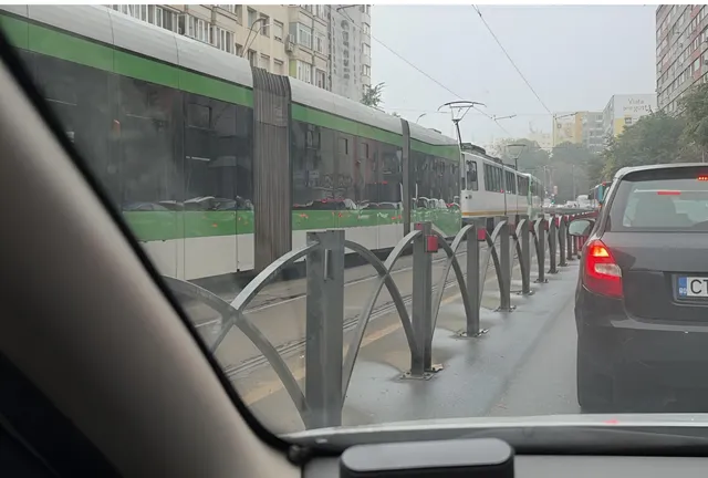 Un tramvai verde și alb circulă pe șine lângă un gard metalic gri și un autoturism gri în partea dreaptă.