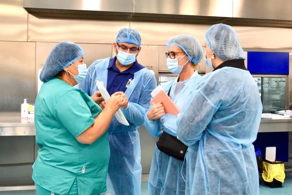 Patru persoane purtând chirurgia, măști și bonete de protecție stau într-o încăpere cu suprafețe metalice și discută ținând în mâini diverse obiecte albe și roz.
