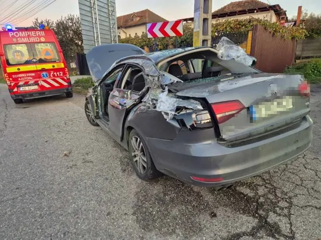 O mașină gri cu capota deschisă, portierele și portbagajul severely damage, staționată pe asfalt lângă o ambulanță roșie.