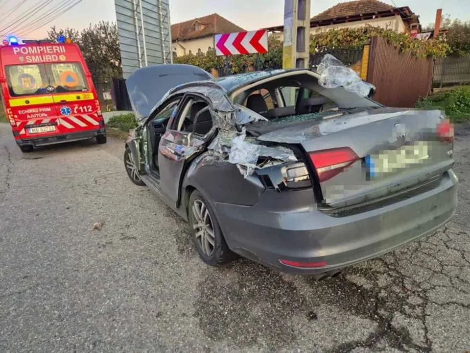 O mașină gri cu capota deschisă, portierele și portbagajul severely damage, staționată pe asfalt lângă o ambulanță roșie.