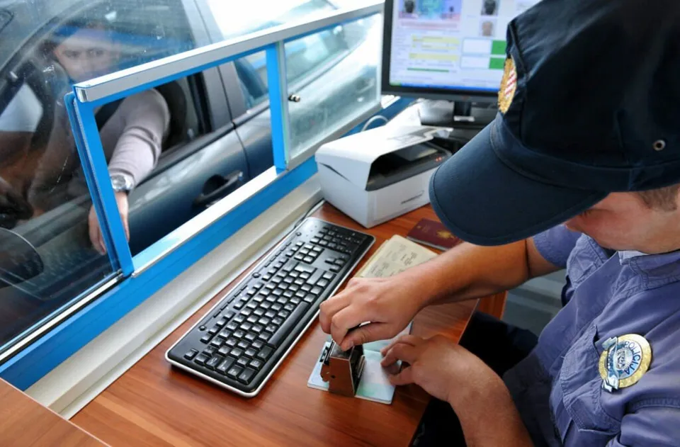 Un agent de poliție stă la un birou lângă un monitor, un tastatură și un pașaport, în timp ce o persoană este vizibilă într-un vehicul.