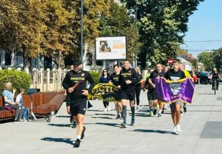 Un grup de bărbați în haine negre ale Invictus aleargă pe un trotuar larg, purtând un steag mov și unul galben.