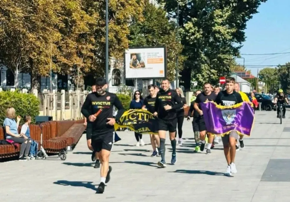Un grup de bărbați în haine negre ale Invictus aleargă pe un trotuar larg, purtând un steag mov și unul galben.