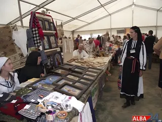 O femeie într-un costum tradițional negru și alb stă lângă un masa lungă cu obiecte artizanale sub un cort alb.