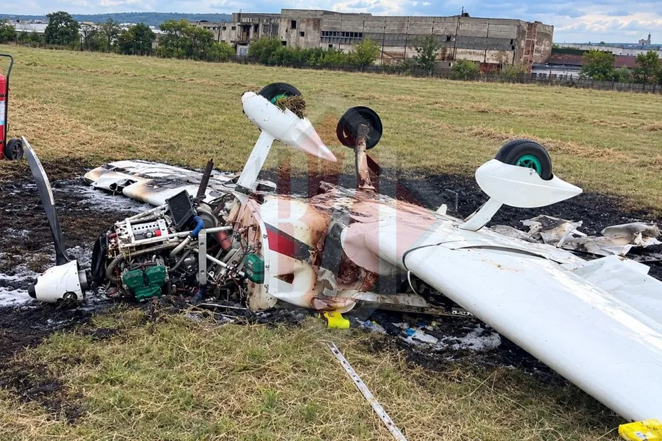 Un avion mic, alb cu detalii roșii, este avariat și ars pe un câmp de iarbă lângă o clădire lungă în fundal.