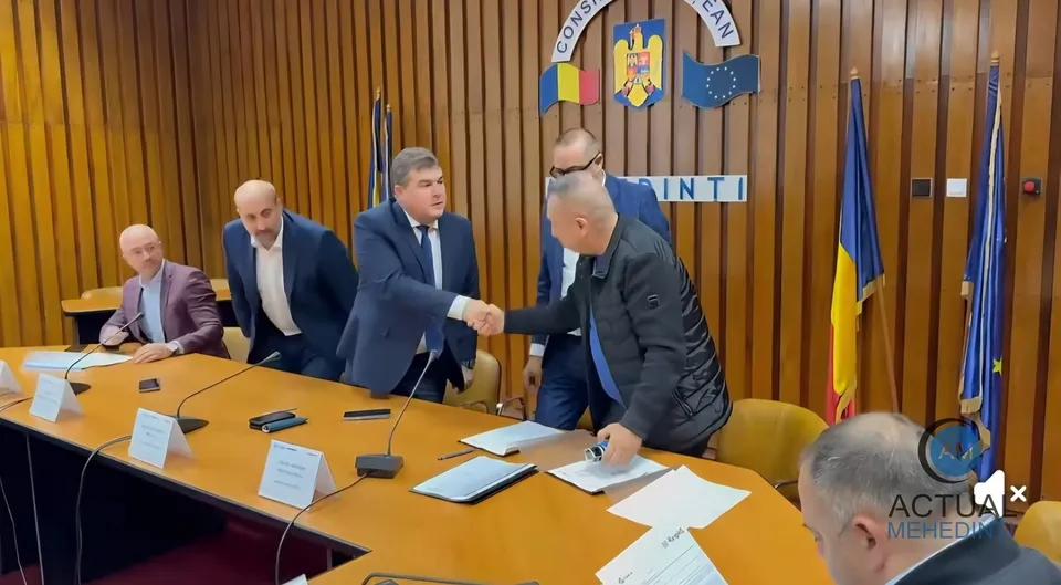 Cinci bărbați stau la o masă lungă de conferințe, doi dintre ei strângând mâna în timp ce restul privesc spre aceștia.