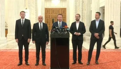 Cinci bărbați în costume scurte stau în picioare în fața unui podium cu microfoane într-o încăpere cu coloane și podea roșie.
