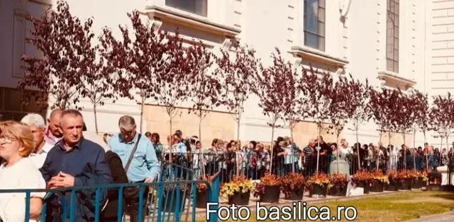 O mulțime de oameni stă în spatele unui gard metalic albastru, lângă un rând de copaci cu frunze purpurii și ghivece cu flori.