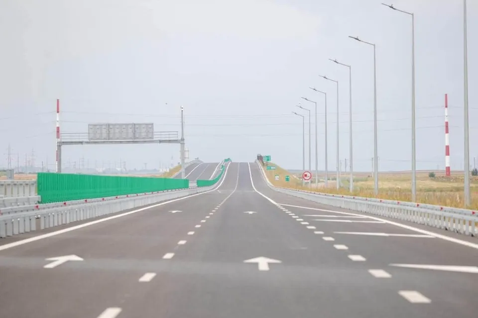 O autostradă cu mai multe benzi de rulare, marcaje albe pe asfalt și stâlpi de iluminat dispuiși pe partea dreaptă sub un cer înnorat.