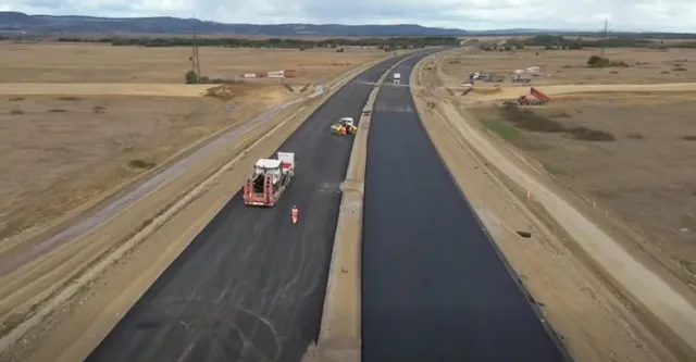 O vedere aeriană prezintă două benzi de asfalt proaspăt turnat, separate de un separator central, înconjurate de terenuri deschise și utilaje de construcție.