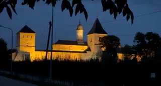 O clădire veche cu turnuri este iluminată nocturn, fiind înconjurată de vegetație sub un cer închis la culoare.